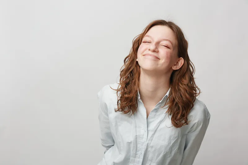 Smiling young woman with curly brown hair wearing white shirt stands against plain background. She looks happy and relaxed with closed eyes and slight head tilt. Positive emotion concept.
