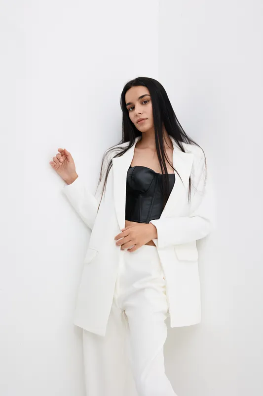young woman wearing black corset and white blazer standing against white wall with long straight dark hair and neutral expression