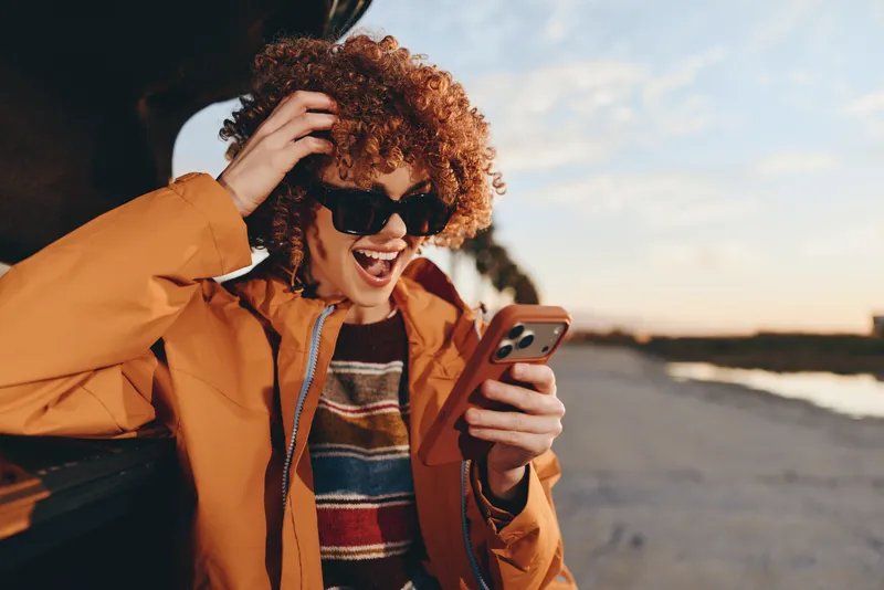 woman lifestyle curly hair laughing as she uses a smartphone on the beach road at sunset, orange jacket, casual travel, bright evening sky