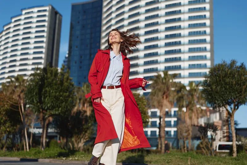 Young woman in red coat walking outdoors near modern high-rise buildings on a sunny day. Casual fashion, urban lifestyle, and confident mood captured in natural light.
