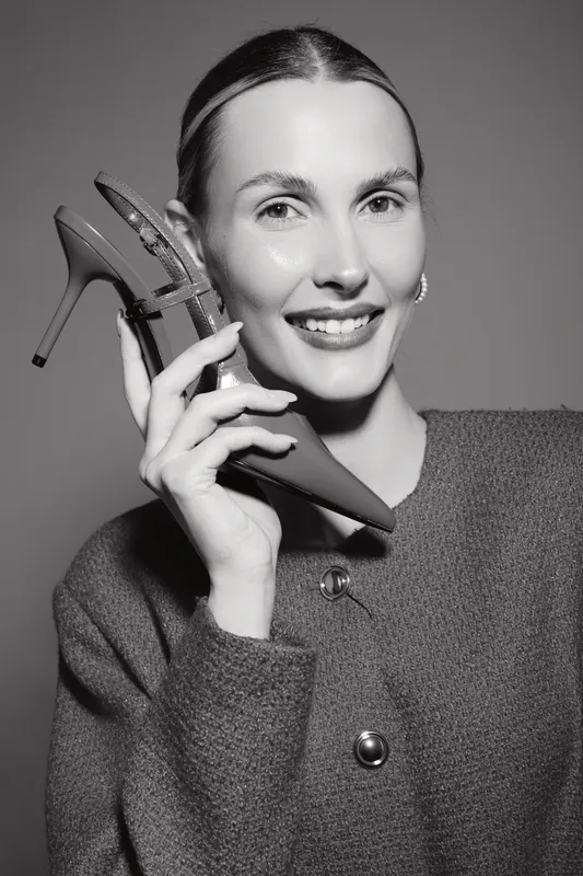 Smiling Caucasian woman with fair skin holds high heel shoe near face, dressed in textured coat. Studio portrait with monochrome lighting, concept for fashion, elegance, and style.