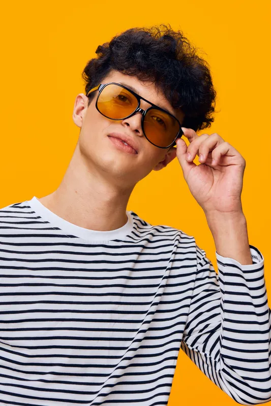 Young man wearing yellow sunglasses and striped shirt poses confidently against vibrant orange background, showcasing modern fashion and casual style with curly hair.