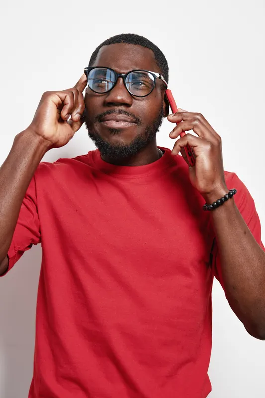 African man with dark skin wearing glasses and red shirt talking on smartphone and thinking with finger on temple isolated on white background. Casual style and focused expression.