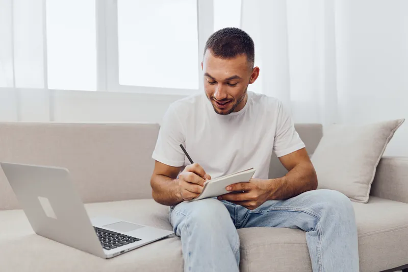 Smiling man writing notes with a laptop nearby, showcasing modern workspace vibes and creativity. Bright and airy setting promotes motivation and productivity.