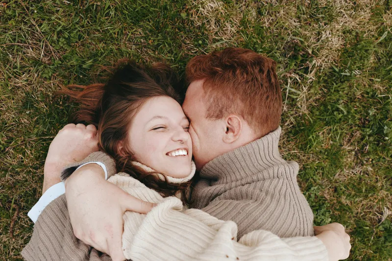 A couple lying on grass in a cozy embrace, smiling with affection, capturing intimate outdoor romance, warm sweaters, natural light, candid moment, relaxed mood, rustic backdrop, lifestyle scene