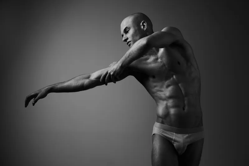 Muscular man striking a pose in a dramatic black and white setting, showcasing strength and expression with intense focus on the body language.