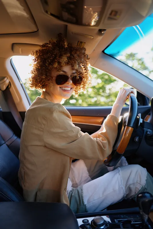 Young woman with curly hair drives a car wearing sunglasses, smiling at the wheel in a warm retro-toned scene that suggests a cheerful, carefree everyday drive.