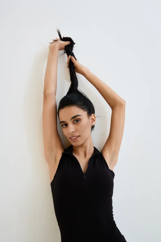 Young woman in black sleeveless outfit tying her long dark hair against white background, showcasing natural beauty and casual style in a minimalist setting.