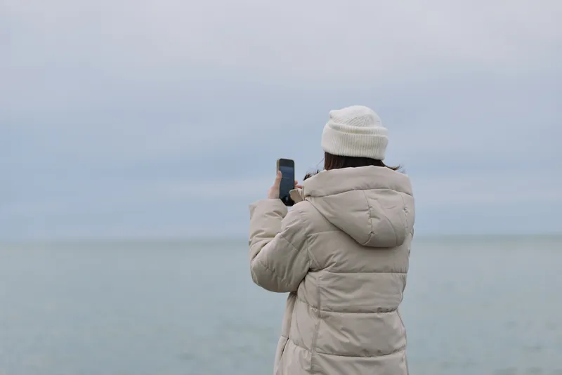 Person in a winter coat and beanie stands by the sea, photographing the coastline with a smartphone, facing calm water under a muted cloudy sky during the day.