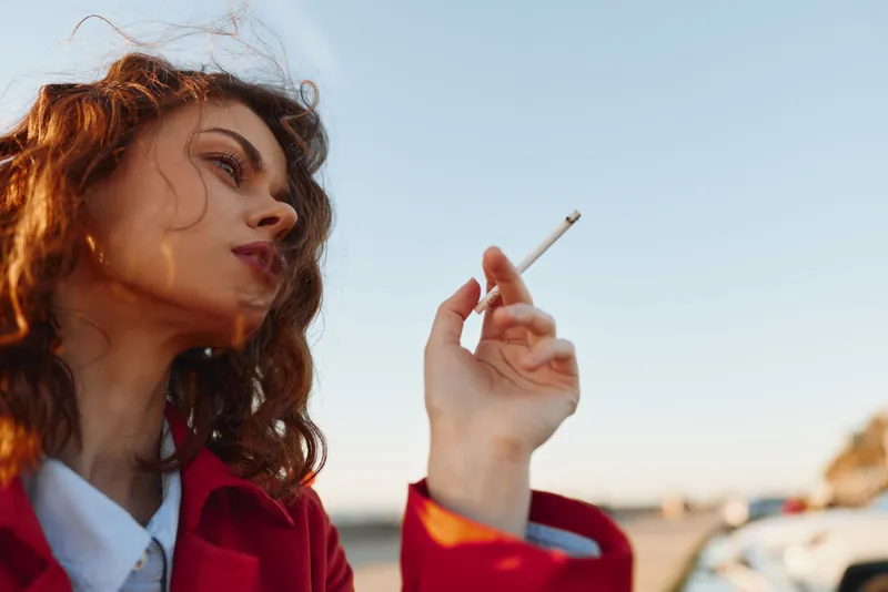 A young woman with curly hair in a red jacket smokes a cigarette outdoors during daytime, looking away thoughtfully. Clear sky and blurred background add depth.