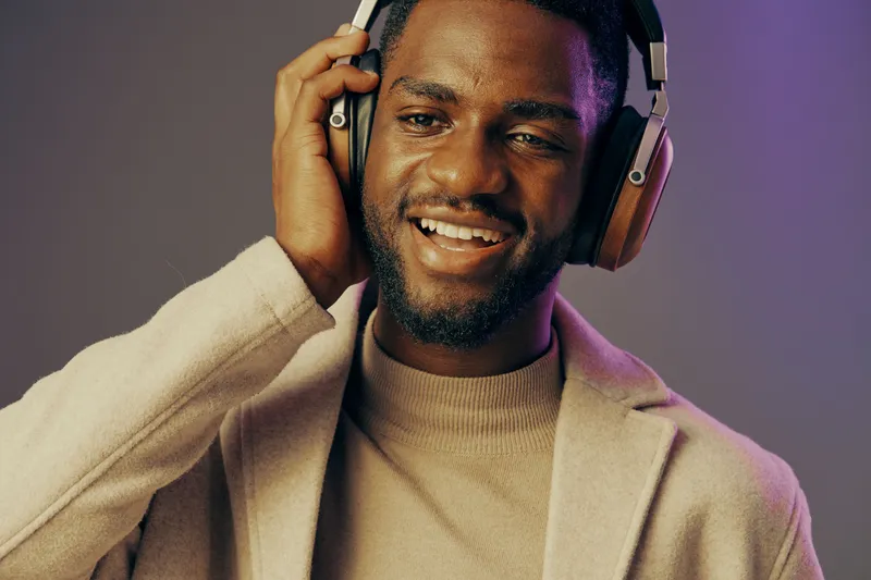 Young man enjoying music with headphones, smiling joyfully against a colorful background, dressed in a beige coat and light turtleneck