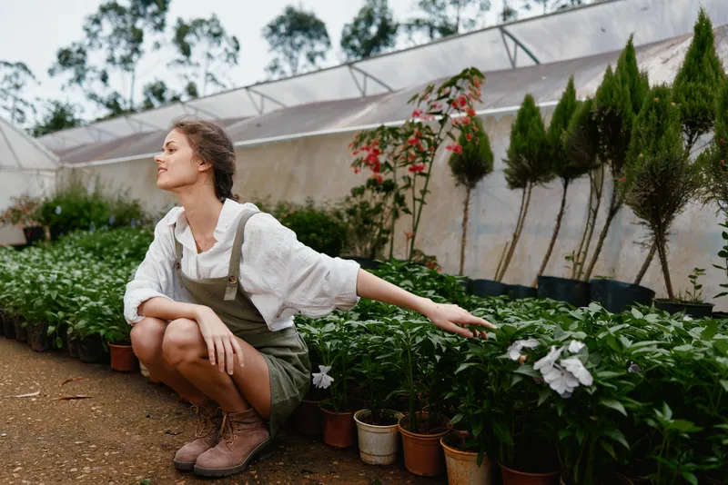 Young woman in a green house touching plants, wearing a casual outfit with a serene expression, surrounded by greenery and colorful flowers