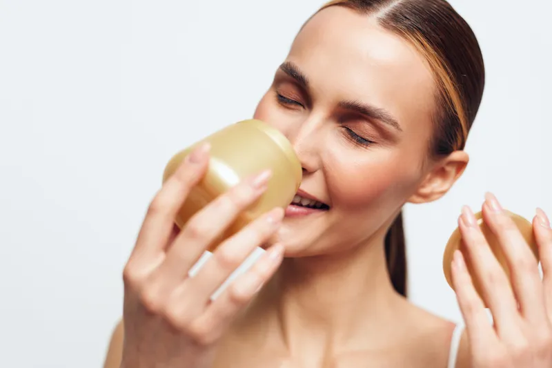 Young woman with smooth skin enjoys the scent of a cream jar while smiling with closed eyes against a plain white background in a skincare concept.
