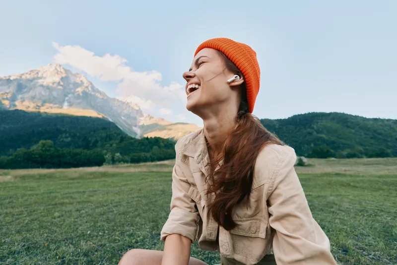 Happy woman wearing orange beanie and wireless earbuds laughing outdoors in nature with mountains and green field in background. Concept of lifestyle and freedom.