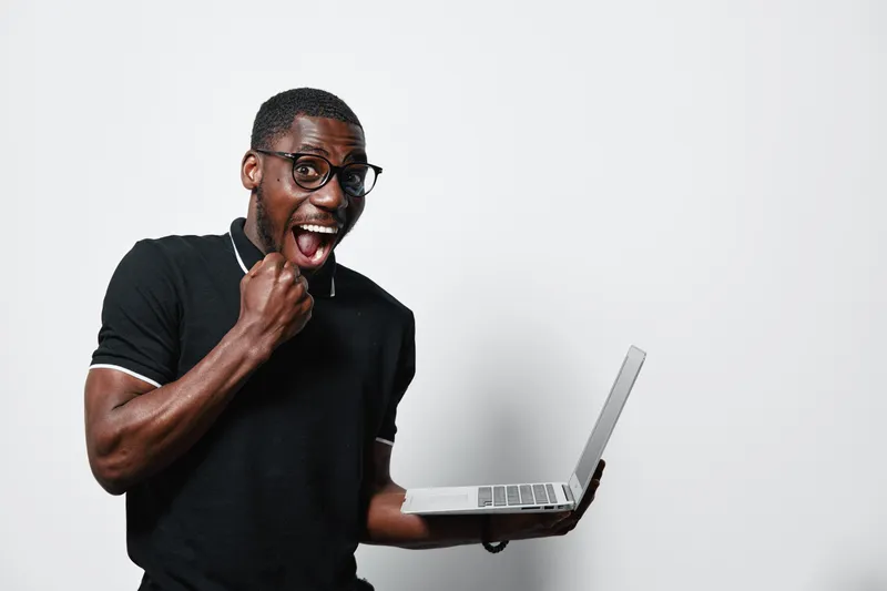 Excited African American man with dark skin and glasses holding laptop and celebrating success with fist pump on white background. Young black male in casual black polo shirt.