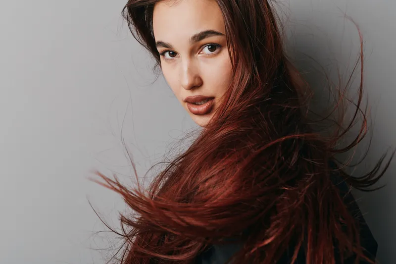Woman with long red hair and natural makeup posing against gray background. Portrait of young female model with flowing hair and serious expression looking at camera.