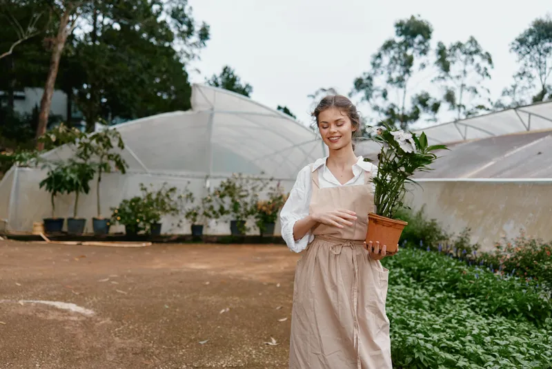 Young woman in a beige apron smiling while holding a potted plant, surrounded by a greenhouse, showcasing a connection with nature and gardening