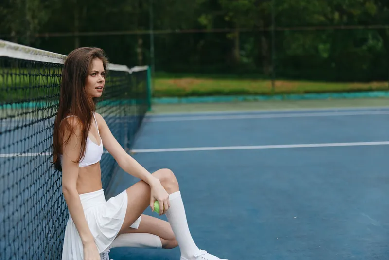 Young athletic woman in sporty outfit sitting on a tennis court, expressing focus and determination, surrounded by a vibrant green background, ideal for fitness themes