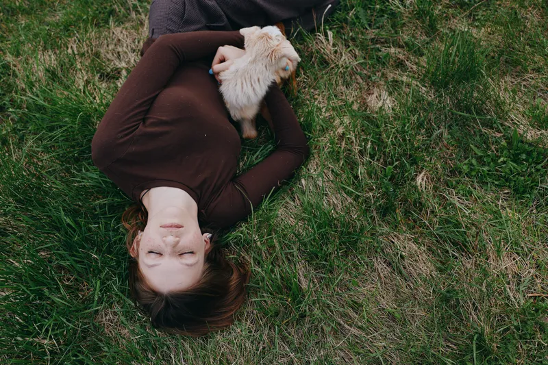 A woman lies on grass in a brown sweater, holding a small fluffy dog close. Natural light highlights a calm, intimate moment of companionship and everyday relaxation.