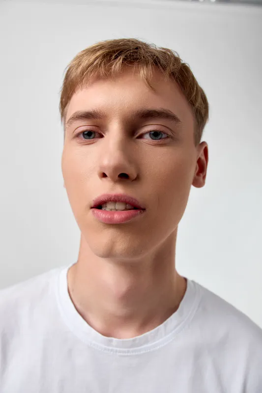 Close-up of a young man with short blond hair, white shirt, neutral expression, looking at the camera with soft light and clean background, suitable for lifestyle, fashion or stock uses