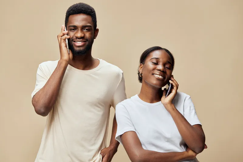 Happy African American couple talking on smartphones, dressed in casual clothing, smiling against a neutral beige background