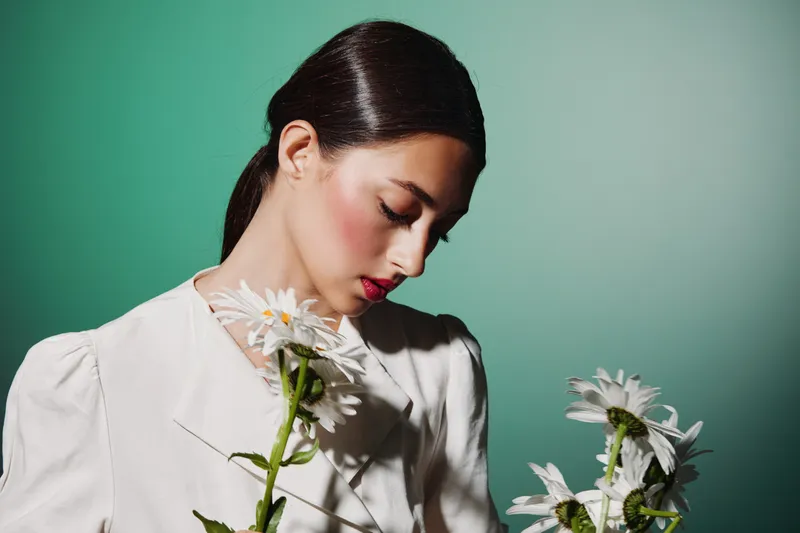Young woman in white fashion attire holds white daisies, posing thoughtfully against a teal background. Concept captures modern beauty, creative styling, and delicate floral elegance.
