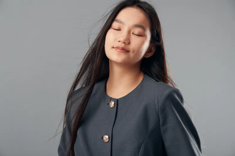 Young woman with long dark hair and closed eyes wearing a dark gray blazer with decorative buttons against gray background. Calm, relaxed female portrait with natural expression.