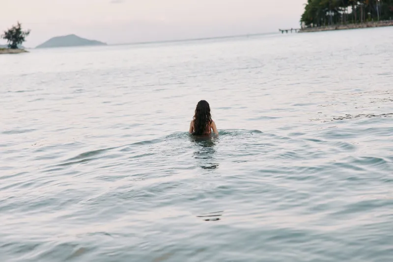 A lone swimmer seen from behind rises from calm sea, soft light on the horizon, distant shore and palm silhouettes, creating a serene, minimal, contemplative mood.