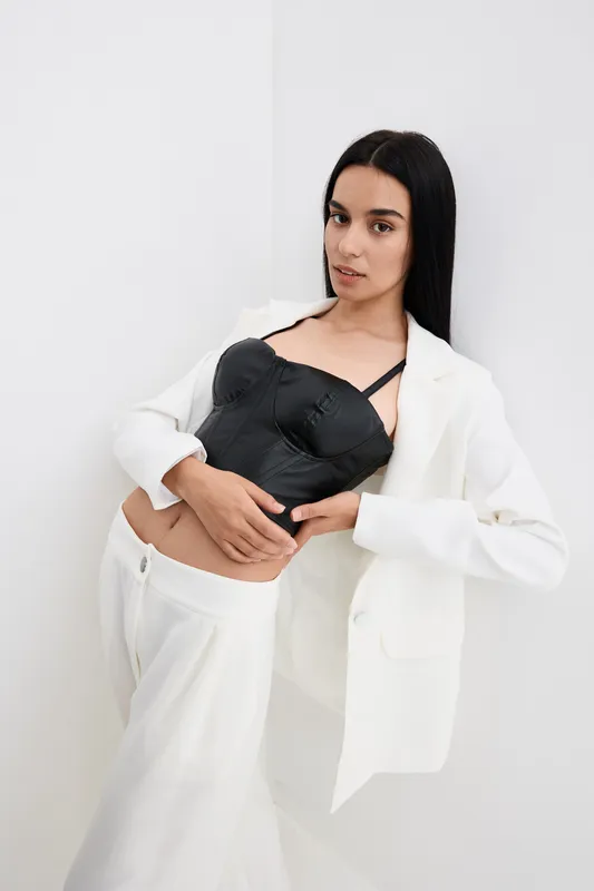 Young woman posing in a stylish black corset and white suit, standing against a plain white background with a confident and poised expression.