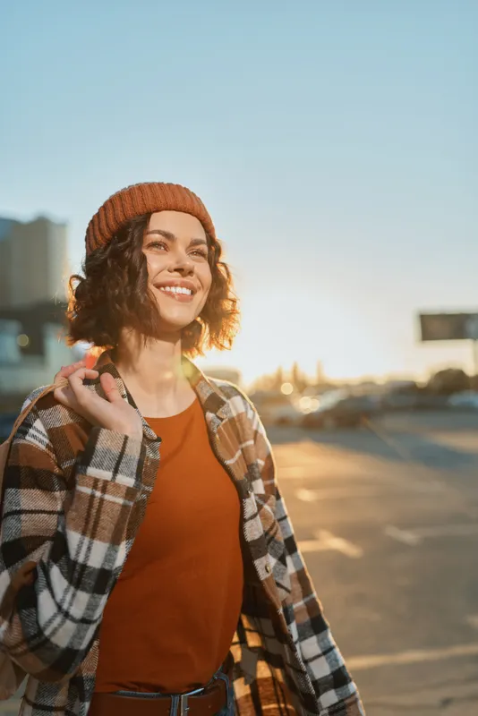 Young woman smile in urban street at golden hour, candid portrait with beanie and plaid coat, warm golden hour glow, authenticity, candid lifestyle, mindful living, emotional storytelling
