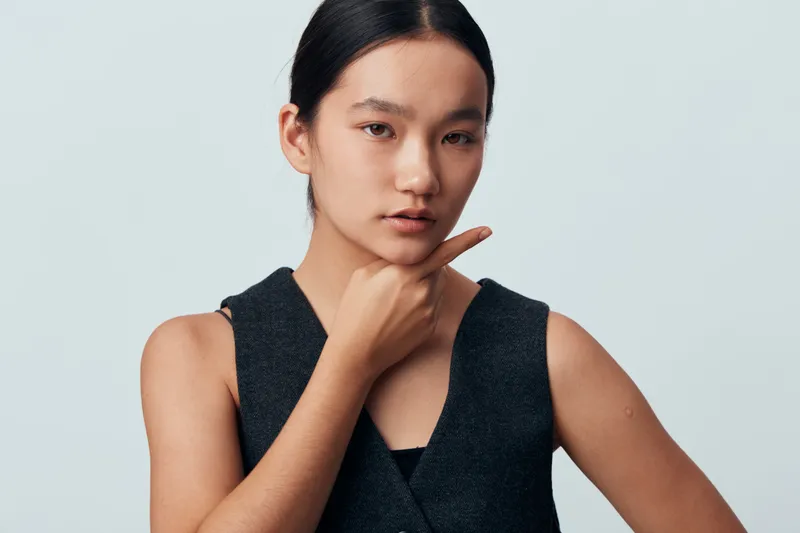 Young woman with thoughtful expression wearing sleeveless dark vest posing with hand near chin on plain light background. Studio portrait with natural makeup and neat hairstyle.