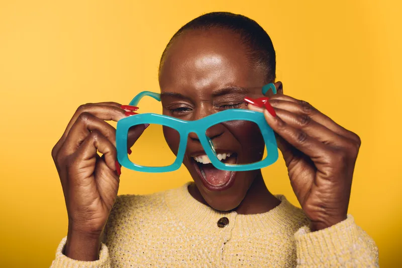 Happy African woman with dark skin wearing yellow sweater holds oversized blue glasses, laughing with eyes closed. Bright yellow background creates joyful and playful mood for lifestyle concept.