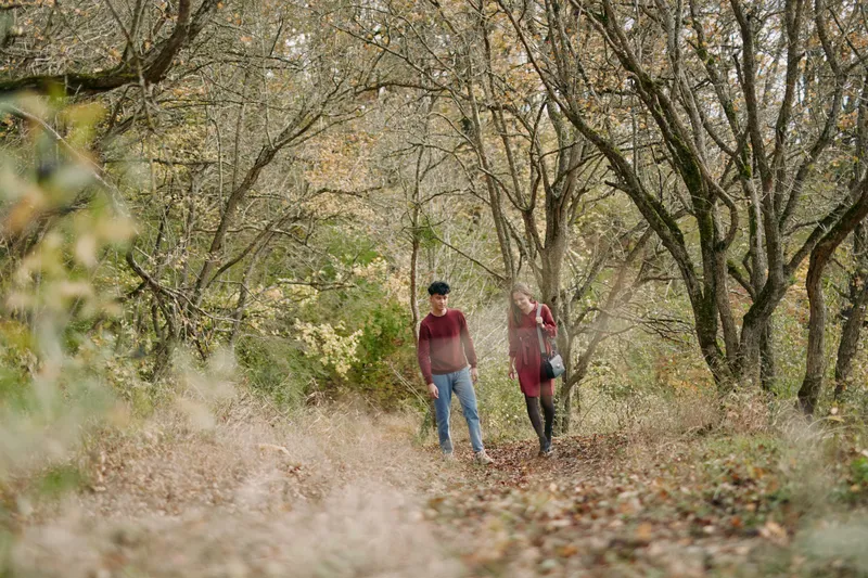 A genuine moment unfolds in a quiet forest, as two people walk a leaf strewn path under soft autumn light, creating a believable and candid scene.
