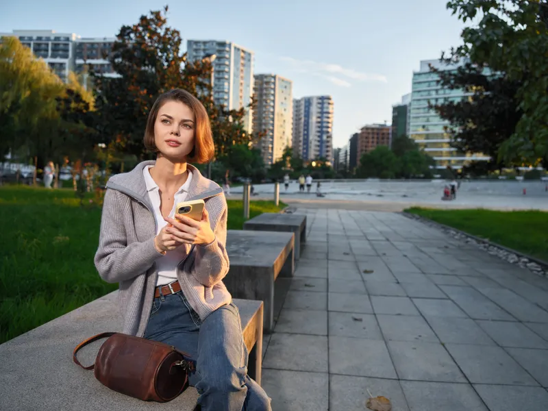 A stylish young woman sits on a concrete bench in an urban park, looking at her phone with a calm expression. Modern buildings rise in the background, creating a relaxed, contemporary vibe and a