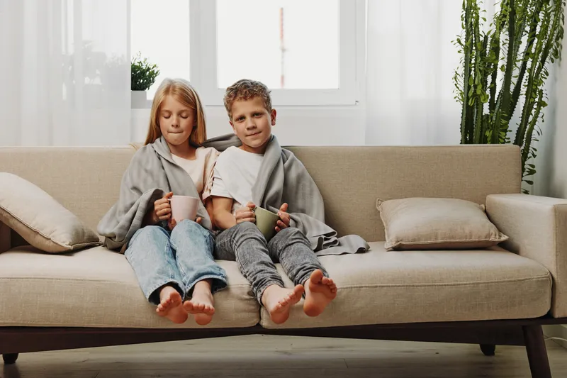 Two children wrapped in cozy blankets sit barefoot on a beige sofa holding warm mugs in a bright living room with plants and natural light. Concept of comfort and family.