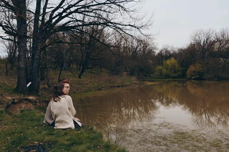 Woman sits by a tranquil lake in a park wearing a cozy sweater, reflecting on a quiet day. Soft light and muted tones create a calm mood, highlighting nature and autumn ambiance.