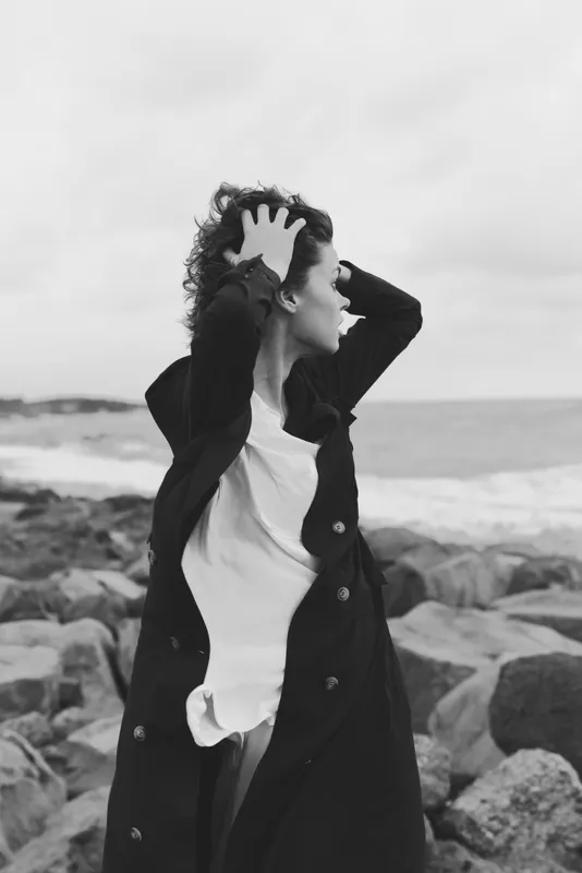 Woman in a long coat stands by a windy coastline, hands through her hair, facing the sea. Monochrome image highlighting mood, resilience, and coastal atmosphere for fashion, lifestyle, or editorial