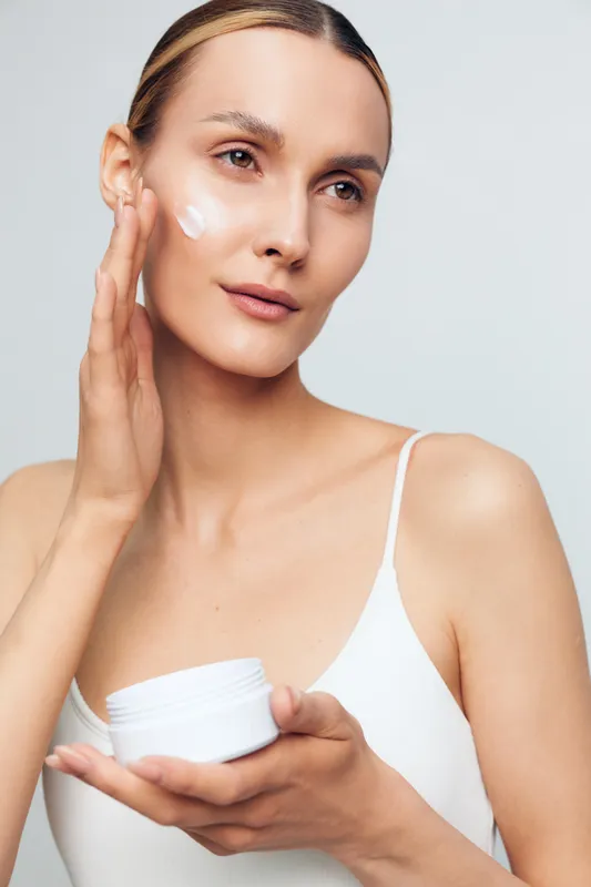 Young woman applies moisturizer to her face, smiling softly as she looks slightly upward, standing against a plain light background, showcasing a calm skincare moment