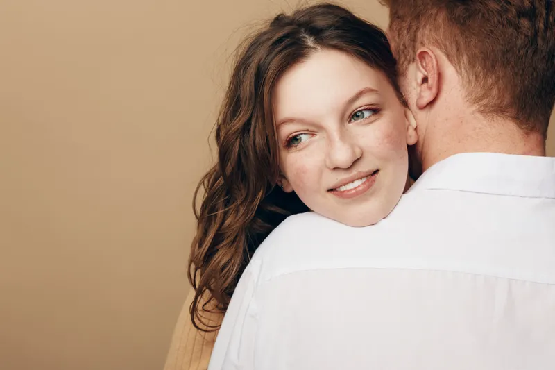 Young woman with curly hair smiles gently while hugging man in white shirt on beige background. Concept of love, affection, relationship, and intimacy between couple.