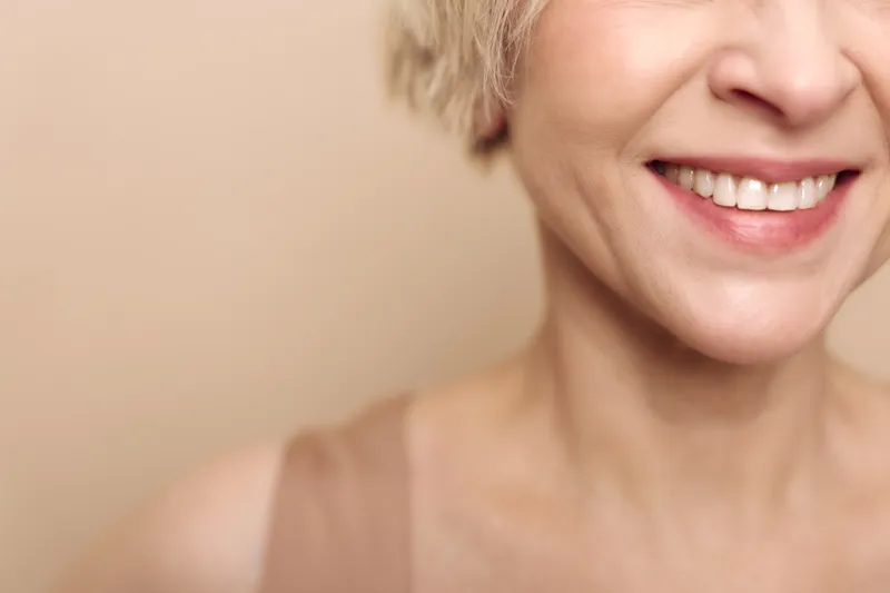 Close-up of smiling woman with short blonde hair shows teeth and lips on beige background. Concept of happiness, beauty, skincare, natural expression, and positive mood.
