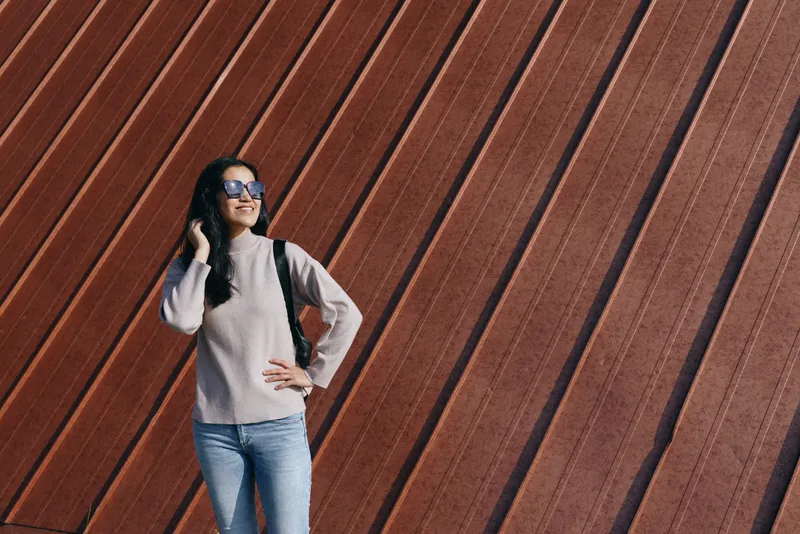 Casual woman with backpack stands beside a diagonal brown wall, wearing sunglasses and a cozy sweater, posing confidently for lifestyle, travel and street photography.