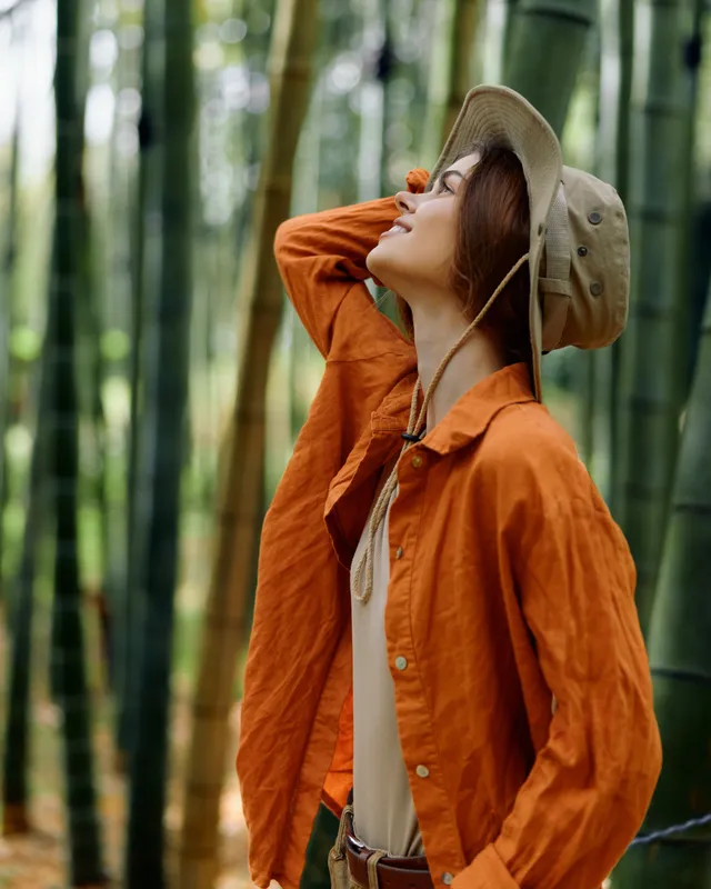 woman in hat and orange shirt stands in bamboo forest, portrait style travel photo of nature and outdoors, female explorer enjoying green trees and sunlight with serene relaxed expression