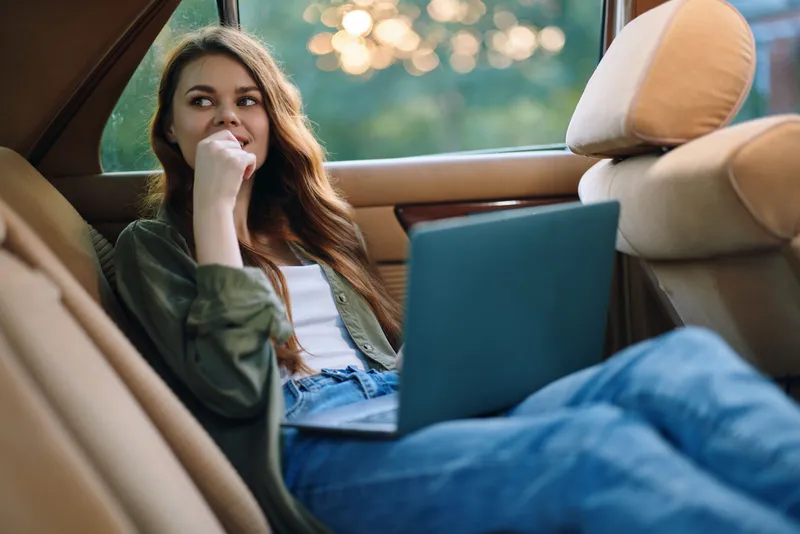 Relaxed young woman working on a laptop in a car, showcasing a casual style with natural light in the background, expressing contemplation and creativity.