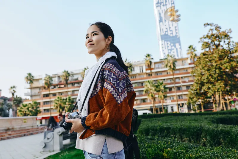 Traveler woman with camera outdoors in urban landscape scene, wearing a scarf and backpack, looking ahead among palm trees and modern buildings, capturing a candid street moment with warm light.