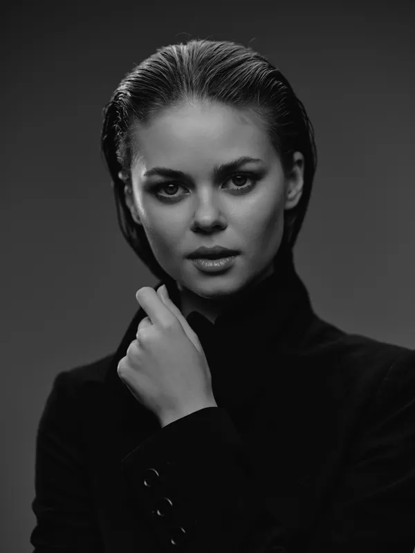 Black and white portrait of a serious woman with slicked-back hair wearing a dark jacket in studio lighting.