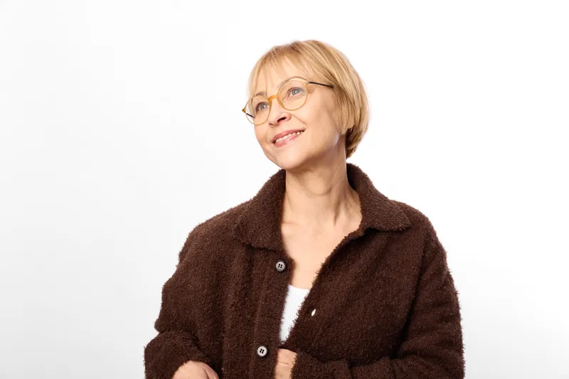 Senior woman portrait with glasses, warm smile, brown coat, indoors headshot, relaxed pose, white background, soft light, studio minimalism, calm expression, friendly demeanor