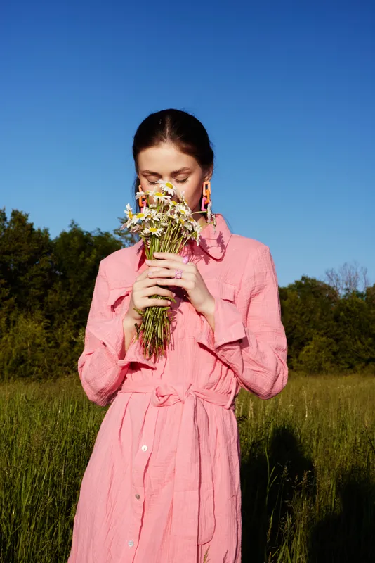 A young woman in a pink dress holds a bouquet of wildflowers, enjoying the beauty of nature in a sunny meadow with a clear blue sky