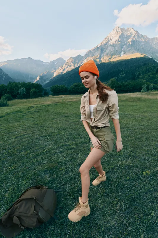 A young woman in casual outdoor clothing and an orange beanie walks on green grass with mountains in the background. Lifestyle concept of adventure and nature exploration.