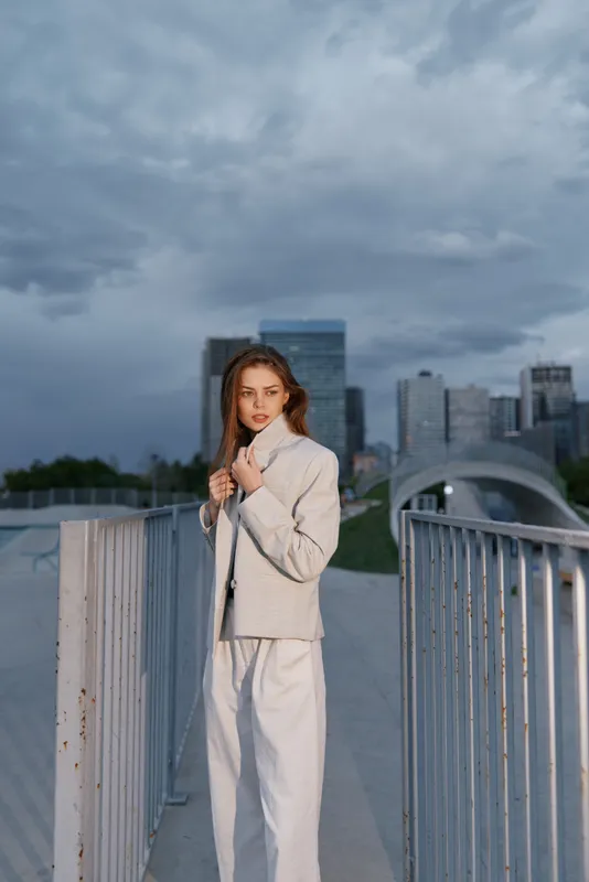 Woman in light beige suit standing on urban bridge under cloudy sky with modern cityscape backdrop, fashion style, confident expression, outdoor business concept