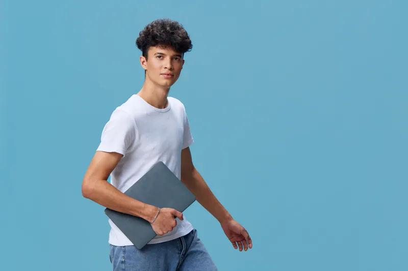 young man holding laptop isolated on blue background, casual clothing, confident expression, curly hair, technology concept